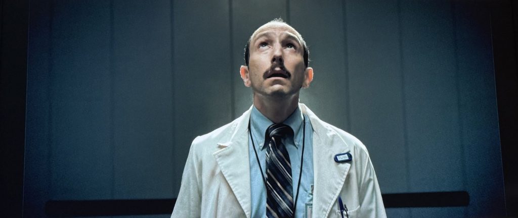 Stranger Things screenshot of a man in a white labcoat looking up while standing inside an elevator