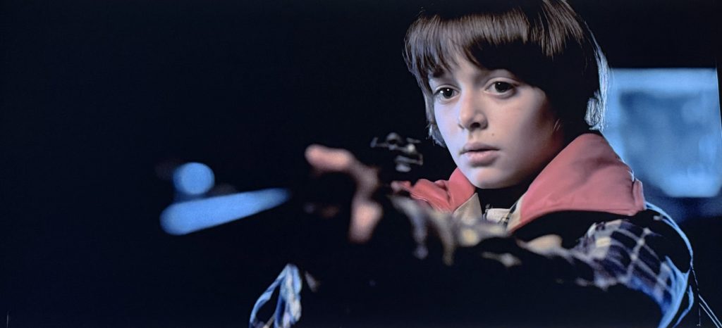 Stranger Things screenshot. Will Byers points a rifle in the direction of the viewer.