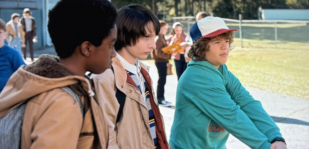 Stranger Things screenshot. Three young boys stand facing two boys who are off-screen. The boy on the left holds his arms out in front of him.