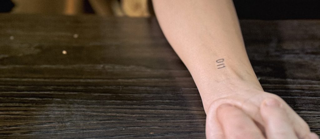 Stranger Things screenshot. Benny examines a young girl's arm, which has a tattoo that reads "011" on her wrist.