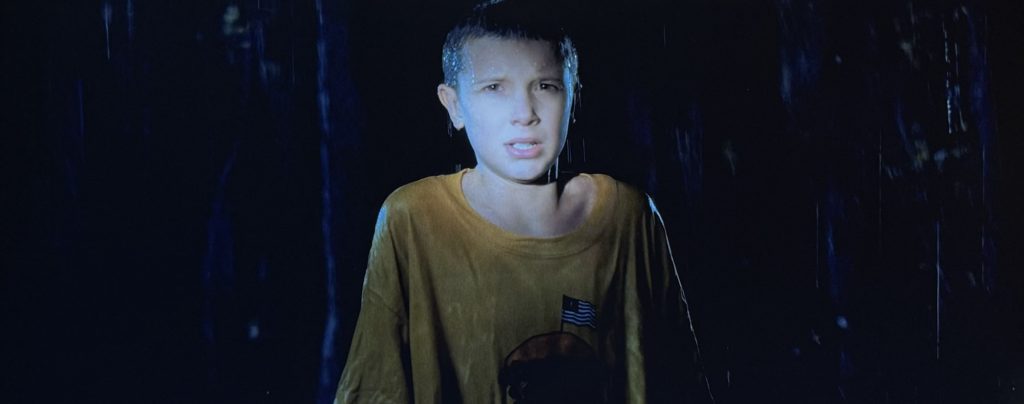 Stranger Things screen shot. Eleven stands in the rain, illuminated by the boys' flashlights.